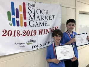 Two students with certificates and Stock Market Game banner